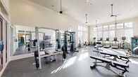 Located at 5771 Oakwood Rd NW in Huntsville, Alabama, this single-floor fitness center offers a spacious and thoughtfully designed workout facility. The space is divided into distinct functional zones, each tailored to a specific type of exercise, all unified by a consistent aesthetic of light-toned walls, crown molding, and durable speckled rubber flooring throughout.

The main workout area is impressively open, with soaring ceilings, recessed lighting, and ceiling fans that contribute to a bright and airy atmosphere. Floor-to-ceiling windows and large multi-pane glass panels line the walls, flooding the space with natural light and providing outdoor views of the surrounding community. A wall-mounted flat-screen television adds convenience for users during their workouts.

The strength training section is well-appointed with a cable crossover machine, a power rack with free weights, a full dumbbell rack with mirror backing, and multiple adjustable benches. The cardio zone is equally comprehensive, featuring treadmills, stationary bikes, elliptical machines, and recumbent bikes positioned to take full advantage of the natural light.

A separate studio room provides a dedicated space for group or individual training, equipped with spin bikes, medicine balls, kettlebells, stability balls, resistance equipment, and mirrored walls. Elegant wood-framed glass doors connect the rooms, maintaining an open and cohesive feel throughout the facility.