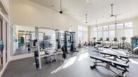 Located at 5771 Oakwood Rd NW in Huntsville, Alabama, this single-floor fitness center offers a spacious and thoughtfully designed workout facility. The space is divided into distinct functional zones, each tailored to a specific type of exercise, all unified by a consistent aesthetic of light-toned walls, crown molding, and durable speckled rubber flooring throughout.

The main workout area is impressively open, with soaring ceilings, recessed lighting, and ceiling fans that contribute to a bright and airy atmosphere. Floor-to-ceiling windows and large multi-pane glass panels line the walls, flooding the space with natural light and providing outdoor views of the surrounding community. A wall-mounted flat-screen television adds convenience for users during their workouts.

The strength training section is well-appointed with a cable crossover machine, a power rack with free weights, a full dumbbell rack with mirror backing, and multiple adjustable benches. The cardio zone is equally comprehensive, featuring treadmills, stationary bikes, elliptical machines, and recumbent bikes positioned to take full advantage of the natural light.

A separate studio room provides a dedicated space for group or individual training, equipped with spin bikes, medicine balls, kettlebells, stability balls, resistance equipment, and mirrored walls. Elegant wood-framed glass doors connect the rooms, maintaining an open and cohesive feel throughout the facility.