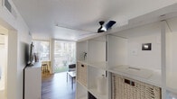 Located at 3130 N 7th Ave in Phoenix, Arizona, this single-floor studio offers a well-organized, open-concept layout that maximizes every square foot. The living area is bright and airy, benefiting from generous natural light streaming through large sliding glass doors fitted with vertical blinds that open to an outdoor patio area with views of lush greenery. Wood-style flooring runs throughout, lending warmth and visual continuity to the space. A sleek open shelving unit with woven basket storage acts as a functional room divider, while a ceiling fan with track lighting overhead ensures year-round comfort and versatility. A curtain partition elegantly separates the sleeping area from the living space, providing privacy without sacrificing the open feel. A skylight adds an additional source of natural light to the interior. The kitchen is efficiently designed, featuring shaker-style cabinetry in soft grey, white subway tile backsplash, white countertops, a stainless steel sink, and a full suite of stainless steel appliances including a range, over-range microwave, and refrigerator. An exposed white brick wall adds a distinctive architectural character. The bathroom includes a full soaking tub and shower combination with chrome fixtures, completing this thoughtfully appointed space.