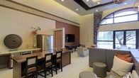 This single-floor space is thoughtfully designed to blend functionality with comfort, offering an open and inviting layout suitable for gathering and relaxation. The kitchenette area features a stainless steel refrigerator, dark wood cabinetry, and a bar-height counter with seating, making it ideal for casual dining or entertaining. Warm earth tones — including rich terracotta and cream-colored walls — are complemented by decorative horizontal banding that adds visual interest and architectural depth. The lounge area is furnished with stylish upholstered seating, including accent chairs and a tufted ottoman, arranged around a wall-mounted flat-screen television, creating a welcoming entertainment hub. A billiards table in the background adds a recreational element to the space. The soaring tray ceiling with recessed lighting enhances the sense of openness and grandeur, while an arched window dressed with patterned drapery floods the space with natural light and hints at lush greenery beyond. The light-toned flooring ties the entire space together, offering a clean and cohesive aesthetic throughout. This space is well-suited as a clubhouse, residents' lounge, or multipurpose amenity area.