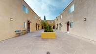 Located in the Van Nuys neighborhood of Los Angeles, this courtyard space serves as the shared entrance and patio area for a multi-unit residential property. The layout is defined by a central open-air courtyard flanked on both sides by two-story stucco buildings finished in a warm sandy beige tone, creating an enclosed yet airy atmosphere characteristic of Southern California architecture.

The patio features a distinctive flagstone-patterned concrete floor that spans the full length of the courtyard, lending a Mediterranean-inspired aesthetic to the space. A large, boldly colored raised planter serves as the visual centerpiece, filled with tropical and drought-tolerant plants including palm trees and various succulents, adding a lush, verdant focal point to the otherwise open expanse.

Individual unit entrances line both sides of the courtyard, each accented with decorative metal-grille security doors and brown awnings that provide a cohesive, structured appearance. Wall-mounted exterior light fixtures are positioned between doorways, ensuring functional illumination throughout. At one end, a covered passageway provides access to and from the street, while open sky overhead fills the courtyard with abundant natural light. A seating area with chairs is also present, further enhancing the communal outdoor ambiance of this inviting shared space.