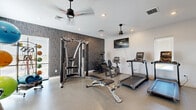 Located in Greenfield, Indiana, this dedicated fitness center offers a thoughtfully designed exercise space equipped for a comprehensive workout experience. The room features durable speckled flooring ideal for high-traffic athletic use, complemented by crisp white walls and a striking textured dark accent wall that adds a modern, energetic aesthetic to the environment.

The space is outfitted with a robust selection of equipment, including multiple Matrix treadmills, a recumbent bike, an elliptical, and a multi-function cable strength-training machine. A adjustable weight bench and a full rack of dumbbells support free-weight training, while medicine balls, stability balls, and additional accessories are neatly organized on a dedicated rack for functional fitness and flexibility work.

A large wall-mounted flat-screen television is positioned for easy viewing during cardio sessions. Full-length mirrors along one wall enhance the sense of openness and allow users to monitor their form during exercise. Two ceiling fans with pendant lighting provide airflow and illumination, keeping the space comfortable during intense workouts. Recessed lighting throughout adds to the bright, clean atmosphere.

Natural light filters in through slatted windows, contributing to an inviting and motivating environment. The overall design is modern and functional, making this fitness center a well-rounded facility for residents seeking convenient, on-site exercise options.