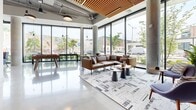 Located at 525 Lincoln St in Boston's Allston neighborhood, this expansive single-floor communal space offers a thoughtfully designed, open-plan layout that blends industrial and contemporary aesthetics. Polished concrete floors reflect abundant natural light streaming through dramatic floor-to-ceiling glass walls, which frame lively street-level views including a colorful neighborhood mural. Exposed ductwork and structural concrete columns contrast warmly with wood-slat ceiling accents and natural wood furnishings throughout.

The space is organized into distinct yet flowing zones. A lounge area features a leather sofa, accent chairs, and a coordinating area rug anchored beneath large corner windows. Nearby, a foosball table adds a playful, social element. A communal dining zone is anchored by a solid wood table surrounded by upholstered blue chairs, positioned beside a vertical wood-slat partition that adds warmth and architectural definition. A kitchenette with light cabinetry and open shelving provides convenient service support.

Elsewhere, colorful lounge seating clusters near additional window walls, while a media nook showcases a textured metallic accent wall with a wall-mounted display. Glass-enclosed private rooms offer quieter spaces within the broader open layout. The entrance features a sleek glass vestibule, seamlessly connecting the interior to the outdoors.