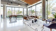 Located at 525 Lincoln St in Boston's Allston neighborhood, this expansive single-floor communal space offers a thoughtfully designed, open-plan layout that blends industrial and contemporary aesthetics. Polished concrete floors reflect abundant natural light streaming through dramatic floor-to-ceiling glass walls, which frame lively street-level views including a colorful neighborhood mural. Exposed ductwork and structural concrete columns contrast warmly with wood-slat ceiling accents and natural wood furnishings throughout.

The space is organized into distinct yet flowing zones. A lounge area features a leather sofa, accent chairs, and a coordinating area rug anchored beneath large corner windows. Nearby, a foosball table adds a playful, social element. A communal dining zone is anchored by a solid wood table surrounded by upholstered blue chairs, positioned beside a vertical wood-slat partition that adds warmth and architectural definition. A kitchenette with light cabinetry and open shelving provides convenient service support.

Elsewhere, colorful lounge seating clusters near additional window walls, while a media nook showcases a textured metallic accent wall with a wall-mounted display. Glass-enclosed private rooms offer quieter spaces within the broader open layout. The entrance features a sleek glass vestibule, seamlessly connecting the interior to the outdoors.