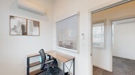 This single-floor professional office suite offers a thoughtfully designed workspace distributed across multiple rooms. The layout begins with a welcoming reception area featuring vaulted ceilings, recessed lighting, and a horizontal window that frames views of surrounding greenery. A white front desk anchors the space, complemented by a warm cognac leather accent chair and a small side table, creating a polished yet approachable entry point.

A central corridor lined with windows fitted with white blinds connects the reception area to several private office rooms. Each individual office is consistently appointed with ergonomic mesh task chairs, wood-and-metal L-shaped or straight desks, wall-mounted mini-split HVAC units for independent climate control, and framed abstract artwork. Natural light filters through the blinds in each room, contributing to a bright and focused work environment. Patterned, textured carpet in a dark neutral tone runs throughout the offices and hallway, adding visual warmth.

The suite also includes an ADA-compliant restroom featuring grab bars, light wood-look luxury vinyl flooring, and practical storage furnishings. Overall, the space presents a clean, modern, and functional professional environment well-suited for focused individual or small-team office use.