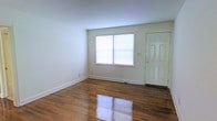 This single-floor space offers a clean, straightforward layout with a welcoming entry area visible upon arrival. The main living area is defined by gleaming, dark-toned hardwood floors that run throughout, adding warmth and character to the otherwise neutral interior. White walls provide a bright, versatile backdrop, while a large window along the front wall allows natural light to fill the room, enhancing the open feel of the space. The paneled entry door, painted white, adds a classic touch to the entryway. Baseboard trim lines the perimeter of the room, contributing to a tidy, well-maintained appearance. The overall ambiance is simple and functional, offering a blank canvas well-suited for a variety of uses and furnishing styles.