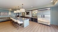 Located in Huntersville, NC, this single-floor community space is thoughtfully designed to serve as a social hub for gatherings and entertainment. The open-concept layout seamlessly connects the kitchen, dining, and lounge areas, all unified by warm hardwood-style flooring and a neutral, sophisticated color palette.

The kitchen is outfitted for serious entertaining, featuring a large central island with a grey stone countertop and seating for multiple guests, illuminated by a trio of elegant drum pendant lights. A commercial-grade stainless steel refrigerator anchors one wall, while dark wood cabinetry and a decorative patterned tile backsplash add visual depth. A large framed mirror reflects the space, enhancing the sense of openness.

The adjacent dining and lounge area provides flexible seating, including a formal dining table set atop a classic area rug, alongside comfortable sofa seating — ideal for both casual mingling and structured gatherings.

A dedicated game room, partially enclosed with refined wainscoting panels painted in a soft sage green, houses a round poker table surrounded by upholstered chairs. Large windows flood this area with natural light and offer views of an outdoor deck. A wall-mounted TV and a dark wood credenza complete the room, making it a versatile and inviting retreat for recreation and socializing.