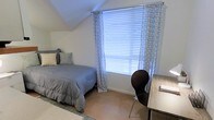This single-floor bedroom features a distinctive angled ceiling that adds architectural character to the space. The room is anchored by a comfortable bed dressed in neutral gray bedding with coordinating pillows, positioned beneath the sloped ceiling for a snug, efficient use of space. A large window serves as the focal point of the room, fitted with horizontal blinds and light patterned curtains that allow natural light to filter in while maintaining privacy. Adjacent to the window, a dark modern chair is paired with a wood-toned desk, creating a functional workspace area. The light-toned flooring complements the white walls, contributing to a clean and airy atmosphere throughout the room. A small area rug adds a touch of softness and warmth to the flooring near the bed. The overall layout is thoughtfully arranged to maximize functionality within the space, making it well-suited for both rest and productivity. The neutral color palette and understated décor give the room a calm, organized ambiance.