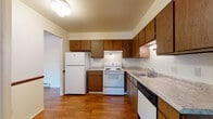 This single-floor, 2-bedroom, 1-bathroom residence in Saginaw, Michigan is thoughtfully laid out to maximize everyday functionality and comfort. The kitchen serves as a practical hub of the home, featuring warm wood cabinetry that extends to the upper walls for generous storage, light granite-style countertops, and a full suite of appliances including a refrigerator, electric range, and dishwasher. Durable hardwood-style flooring runs throughout the kitchen, adding warmth and easy maintenance to the space. The living area is carpeted in a cool blue-grey tone and opens directly to a private outdoor space through a sliding glass door dressed with vertical blinds, allowing for natural light and outdoor access. A wall-mounted air conditioning unit and baseboard heating ensure year-round comfort. Both bedrooms are carpeted and feature ceiling fans with integrated lighting, vertical-blind-covered windows that invite natural light, and baseboard heating. The full bathroom includes a classic white-tiled shower and tub combination with hardwood-style flooring. The overall layout flows seamlessly between the kitchen, living, and bedroom areas, creating a cohesive and practical living environment well-suited for daily life.