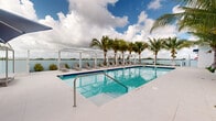 This stunning swimming pool complex in North Miami, FL, exemplifies luxury and tropical charm. The centerpiece is a large rectangular pool with crystal-clear turquoise water, surrounded by a spacious white concrete deck. Palm trees line the perimeter, providing natural shade and a lush, resort-like atmosphere. 

The pool area boasts several amenities, including comfortable lounge chairs for sunbathing and relaxation. A white pergola structure offers a shaded seating area with panoramic views of the adjacent waterway, creating a perfect spot for socializing or quiet contemplation.

The facility adheres to safety standards, as evidenced by the prominently displayed pool rules sign and the presence of life-saving equipment. The architecture of the surrounding buildings features clean, modern lines with white exteriors and blue accents, complementing the overall aesthetic.

Perhaps the most striking feature is the breathtaking waterfront location. The pool area overlooks a serene body of water, with distant views of the Miami skyline, seamlessly blending urban sophistication with natural beauty. This harmonious integration of water elements - from the pool to the scenic waterway - creates a truly immersive aquatic experience.

The meticulous landscaping, featuring a variety of tropical plants and well-manicured green spaces, enhances the sense of a private paradise. This pool area not only provides a space for swimming and sunbathing but also serves as a picturesque retreat that captures the essence of South Florida's coastal charm.