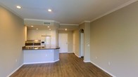 This single-floor residence offers a thoughtfully designed open-concept layout that seamlessly connects the kitchen and living areas, creating a spacious and functional environment. The kitchen is well-equipped with white cabinetry, a stainless steel refrigerator, a built-in microwave, and a curved peninsula counter with a dark countertop, providing ample prep space and a natural divider between the kitchen and the main living area. A sink is positioned along the peninsula, adding to the kitchen's practicality. Recessed lighting in the kitchen area ensures bright, even illumination. Warm hardwood-style flooring flows throughout the space, adding a sense of warmth and cohesion. Crown molding accents the ceilings, lending a refined, classic touch to the interior. The neutral warm-beige wall tones create a welcoming ambiance that complements the flooring and cabinetry. An elegant arched architectural detail adds character near a hallway leading to additional areas of the residence. The entryway is clearly defined with a solid front door, and a thermostat panel is conveniently positioned along the wall. Overall, the space balances modern conveniences with timeless design elements, making it a comfortable and inviting home.