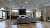 This single-floor space makes a strong architectural impression with its standout coffered ceiling, which adds depth and a sense of refinement to the main lounge area. Recessed lighting is thoughtfully integrated throughout the ceiling, ensuring bright, even illumination across the space. The focal point of the room is a sleek fireplace framed by a rich, dark wood surround, above which a wall-mounted flat-screen television is prominently positioned. Flanking the fireplace are transom windows that allow natural light to filter in, enhancing the open and airy ambiance. The flooring features a striking parquet-style hardwood pattern, lending warmth and visual interest underfoot. Generous sectional seating is arranged around a patterned area rug, creating a comfortable and welcoming gathering area. Neutral-toned accent chairs and side tables are positioned throughout, offering additional seating options. Built-in shelving and a dark wood credenza along the walls provide practical storage while contributing to the space's polished aesthetic. Tasteful wall art adds color and personality to the otherwise neutral palette. Overall, the space balances classic architectural details with contemporary furnishings to create a sophisticated and functional environment.