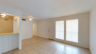 This single-floor space offers a clean, functional layout with a welcoming and airy atmosphere throughout. The main living area is finished with large, light-toned ceramic floor tiles that reflect natural light and lend a sense of openness to the room. A ceiling fan is centrally mounted overhead, providing both practical ventilation and a simple decorative touch.

Generous windows dressed with horizontal blinds line one wall, allowing abundant natural light to pour into the space while maintaining privacy. A front entry door is positioned alongside the windows, creating a bright and accessible entryway. The walls are finished in a crisp white, contributing to the overall bright and neutral palette that makes the space feel open and versatile.

A textured ceiling adds subtle architectural character, while a central hallway leads to the additional rooms, suggesting a straightforward and efficient floor plan. The HVAC vent visible near the ceiling indicates a built-in climate control system for year-round comfort. The overall ambiance is clean, minimal, and move-in ready, offering a neutral canvas well-suited for a variety of interior styles and uses.