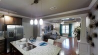 This single-floor space showcases an open-concept design that seamlessly connects the kitchen, dining, and living areas. The kitchen is a standout feature, equipped with a double-basin stainless steel sink, granite countertops with subtle veining, and a trio of pendant lights that illuminate the space with a warm, inviting glow. The countertop extends to serve as a breakfast bar, offering casual seating with white chairs.

The living area is anchored by rich hardwood flooring and a plush area rug, creating a comfortable and defined seating zone. A tray ceiling with crown molding adds architectural depth overhead, while a ceiling fan ensures year-round comfort. Natural light filters through windows dressed with teal curtains, adding a vibrant pop of color against the neutral gray walls.

Decorative wall art and coordinating teal accents throughout the space create a cohesive and stylish aesthetic. The entryway, visible from the kitchen, features a solid door, providing a clear sense of the space's functional layout. Overall, the open floor plan and thoughtful design details combine to create an airy, modern living environment.