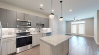 This thoughtfully designed 1-bedroom, 1-bathroom unit offers a contemporary open-concept layout that maximizes functionality and style. The kitchen serves as the heart of the space, featuring sleek gray flat-front cabinetry, a tiled backsplash, granite countertops, and a full suite of stainless steel appliances including a refrigerator, range, microwave, and dishwasher. A central kitchen island with matching granite top and pendant globe lighting provides additional prep space and casual seating. Wood-look flooring flows seamlessly through the kitchen and into the open living area, which benefits from natural light through a set of glass double doors fitted with blinds and a ceiling fan overhead. A dedicated laundry area with a full-size washer and dryer is conveniently located near the kitchen. The bedroom is carpeted for comfort, includes a ceiling fan, and receives natural light through a window. It connects to a spacious walk-in closet outfitted with wire shelving systems for organized storage. The bathroom is well-appointed with a granite-topped double vanity, gray cabinetry with brushed nickel hardware, a large mirror, and a tiled soaking tub-shower combination. Neutral wall tones and clean architectural lines contribute to a cohesive, modern aesthetic throughout.