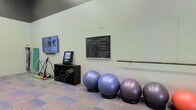 This single-floor fitness space is designed to support a variety of workout styles in a clean, functional environment. The room features light-colored walls and patterned carpet flooring, contributing to a bright and approachable atmosphere. A wall-mounted flat-screen television is positioned for easy viewing during workouts, alongside a chalkboard displaying structured workout routines, making it ideal for group or self-guided exercise sessions. A low storage console unit provides organized space for rolled fitness mats and accessories. Several stability exercise balls are neatly lined along the wall on dedicated holders, offering readily accessible equipment for core and balance training. A horizontal ballet barre is mounted along one wall, supporting stretching, balance, and barre-style exercises. A kiosk-style digital display station is also present, likely providing instructional or scheduling information. The high ceiling adds a sense of openness to the space, preventing any feeling of confinement during active use. Overall, the fitness area is well-organized and versatile, catering to a range of exercise needs from strength training to flexibility and cardio workouts.