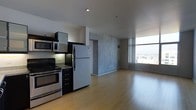 This single-floor space showcases a contemporary open-concept layout that seamlessly blends the kitchen and living areas into one cohesive, light-filled environment. The kitchen is fully equipped with stainless steel appliances, including a gas range with overhead microwave, a full-size refrigerator, and granite countertops, all complemented by sleek dark cabinetry that adds a modern contrast to the space. Warm-toned hardwood flooring extends throughout the entire unit, lending a sense of continuity and comfort underfoot. A striking exposed concrete accent wall serves as an architectural focal point, adding an industrial-chic character that balances the otherwise clean, minimalist aesthetic. Recessed lighting in the white ceiling provides even, ambient illumination throughout. The living area is generously proportioned, offering ample room for furnishing and entertaining. Floor-to-ceiling windows flood the space with abundant natural light and frame sweeping city views, creating a dynamic and energizing atmosphere. The overall ambiance is urban and sophisticated, combining raw industrial elements with polished modern finishes for a stylish and functional living environment.