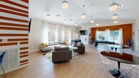 Located on Village Walk Lane in Clemson, South Carolina, this single-floor community clubhouse is designed to foster social interaction and leisure. The space is characterized by an open, airy layout with light hardwood flooring throughout and contemporary globe pendant lighting that adds a modern touch to the interior.

The main lounge area offers ample seating with plush sofas and accent chairs arranged around a central coffee table, all anchored by a colorful area rug. Large windows dressed with sheer white curtains flood the space with abundant natural light, while a wall-mounted flat-screen TV and a bold orange-and-white striped accent wall add visual interest and energy to the room.

Transitioning seamlessly into the game room, residents will find a pool table and a foosball table, catering to a variety of recreational interests. Multiple sets of glass French doors line the rear wall, offering direct views and access to an outdoor patio area, further enhancing the connection between indoor and outdoor living.

A dedicated kitchenette area features a striking deep-blue subway tile backsplash, white quartz countertops, stainless steel appliances, a beverage refrigerator, and a coffee station — making it an ideal spot for casual gatherings. The dining area nearby includes sleek round tables with modern acrylic chairs, completing this well-appointed community amenity space.