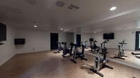 Located at 1668 N St Andrews Place in Los Angeles, this single-floor fitness studio is purpose-built for indoor cycling and group exercise. The space is designed with a clean, modern aesthetic, featuring crisp white walls, light wood-style flooring, and recessed lighting that provides consistent illumination throughout.

The main area of the studio houses multiple stationary spin bikes arranged to accommodate group classes. A full wall of floor-to-ceiling mirrors runs along one side of the room, visually expanding the space while allowing participants to monitor their form during workouts. A textured concrete accent column adds an industrial design element that contrasts with the otherwise minimalist interior.

Wall-mounted flat-screen televisions are positioned at multiple points throughout the studio, enabling instructors to display workout content or metrics for participants. The room offers ample open floor space beyond the bike arrangement, making it adaptable for additional fitness activities or stretching.

Black-framed doors provide access to adjoining areas, and the studio benefits from a glass-panel entry door that introduces a degree of natural light. The overall ambiance is functional yet polished, creating a focused environment well-suited for high-energy indoor cycling sessions in the heart of Hollywood.
