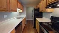 This single-floor residence offers a functional and straightforward layout with a neutral color palette throughout. The galley-style kitchen is equipped with warm oak cabinetry, light countertops, dark hardwood-style flooring, and black appliances including a refrigerator and range, complemented by a stainless steel sink and dishwasher. A dedicated laundry area features tile flooring and an overhead shelf for storage.

The living area is generously proportioned, featuring plush carpeting, baseboard heating, and a large sliding glass door dressed with vertical blinds that allows abundant natural light to fill the room. An adjoining area with hardwood-style flooring includes a ceiling fan, adding versatility to the layout.

The bedrooms are carpeted and each features closet storage, with one room offering a sliding-door closet and a window with vertical blinds. Additional bedroom space includes a walk-in closet with built-in shelving and warm interior lighting.

The full bathroom is well-appointed with an oak vanity, light countertop, a large wall mirror, mosaic tile flooring, and a tub-shower combination with sliding glass doors. Neutral taupe wall tones and white trim carry consistently throughout the residence, creating a cohesive and clean overall ambiance.