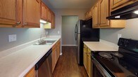 This single-floor residence offers a functional and straightforward layout with a neutral color palette throughout. The galley-style kitchen is equipped with warm oak cabinetry, light countertops, dark hardwood-style flooring, and black appliances including a refrigerator and range, complemented by a stainless steel sink and dishwasher. A dedicated laundry area features tile flooring and an overhead shelf for storage.

The living area is generously proportioned, featuring plush carpeting, baseboard heating, and a large sliding glass door dressed with vertical blinds that allows abundant natural light to fill the room. An adjoining area with hardwood-style flooring includes a ceiling fan, adding versatility to the layout.

The bedrooms are carpeted and each features closet storage, with one room offering a sliding-door closet and a window with vertical blinds. Additional bedroom space includes a walk-in closet with built-in shelving and warm interior lighting.

The full bathroom is well-appointed with an oak vanity, light countertop, a large wall mirror, mosaic tile flooring, and a tub-shower combination with sliding glass doors. Neutral taupe wall tones and white trim carry consistently throughout the residence, creating a cohesive and clean overall ambiance.