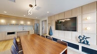 Located at 8460 Ash St in Vancouver, BC, this contemporary single-floor amenity space offers a well-appointed, thoughtfully designed environment ideal for meetings, gatherings, and collaborative work. The space is anchored by a generous conference and dining area centered around a sleek walnut-toned table accommodating multiple guests, surrounded by upholstered chairs and topped by a sculptural pendant light fixture. A floor-to-ceiling built-in wall unit in light wood houses a large display screen, decorative shelving, and woven-panel accents, adding both function and visual warmth. Expansive glazed doors and large windows flood the space with natural light while offering views of the surrounding Vancouver cityscape. A well-equipped kitchenette features light wood cabinetry, a white tile backsplash, open shelving stocked with glassware and dishware, a stainless steel refrigerator, microwave, and sink — all finished in a clean, minimalist style. A window-side counter with amber bar stools provides an additional casual seating area bathed in natural light. A separate lounge nook offers a plush blue armchair beside a large window, complemented by curated artwork. Light hardwood flooring runs throughout, unifying the space with a warm, cohesive aesthetic.