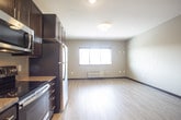 This single-floor unit is designed with a clean, contemporary aesthetic that balances functionality and comfort throughout its layout. The open-concept living and kitchen area serves as the heart of the space, where light wood-tone flooring extends seamlessly underfoot, creating a warm and cohesive atmosphere. Neutral taupe walls complement the overall palette and provide a versatile backdrop.

The kitchen is outfitted with rich, dark espresso cabinetry featuring brushed nickel hardware, granite countertops, and a full suite of stainless steel appliances, including a refrigerator, electric range, and over-the-range microwave. Recessed lighting illuminates the workspace efficiently. The kitchen flows naturally into the living area, which is generously sized and benefits from a window that allows natural light to filter in.

The bathroom continues the design language of the unit, featuring a granite-topped vanity with dark cabinetry, an undermount sink with a chrome faucet, and a large mirror above. A tub and shower combination and chrome towel bar complete the space. Throughout the unit, dark-framed doors add a modern contrast to the light walls and flooring, contributing to a polished, cohesive interior finish.