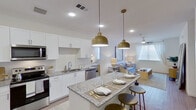 This single-floor residence showcases a thoughtfully designed open-concept layout that seamlessly connects the kitchen, dining, and living areas. The kitchen is a standout feature, equipped with stainless steel appliances including a refrigerator, range, microwave, and dishwasher. Crisp white cabinetry pairs elegantly with granite countertops, while a central island with bar seating serves as both a functional workspace and casual dining spot. A pair of gold dome pendant lights adds a warm, contemporary accent overhead. The living room flows naturally from the kitchen and dining space, bathed in natural light from multiple windows dressed with sheer curtains and blinds. A ceiling fan provides comfort, and the warm hardwood-style flooring ties the open areas together cohesively. The two bedrooms offer a calm, comfortable retreat, each featuring plush carpet, natural light, and ceiling fans or overhead lighting. One bedroom includes a walk-in closet with shelving and a hanging rod. The bathroom is clean and modern, featuring granite-topped vanity cabinetry, a large mirror, and wood-look flooring. Throughout the residence, the bright white walls and consistent finishes create a cohesive, contemporary aesthetic that is both practical and inviting.