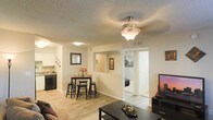 This single-story space showcases an open-concept design that seamlessly connects the living room, dining area, and kitchen. Light wood-style flooring flows throughout, creating a cohesive and welcoming feel. The living area is anchored by a ceiling fan with a light fixture, while a textured ceiling adds subtle character overhead. The dining space comfortably accommodates a bistro-style table with bar stools, positioned adjacent to the kitchen for convenient everyday use. The kitchen features white cabinetry, dark appliances, and a pass-through opening that enhances the sense of openness between spaces. Neutral wall tones provide a versatile backdrop throughout. A hallway visible from the main living area leads to a bedroom, suggesting a well-organized floor plan. Wall-mounted décor and framed artwork add personality without overwhelming the space. The layout is practical and efficient, making the most of the available footprint while maintaining an airy, connected atmosphere suited for comfortable day-to-day living.