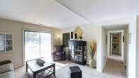This single-floor space centers around a comfortable living room that immediately draws attention with its generous natural light. A full-length sliding glass door, dressed with vertical blinds, allows abundant daylight to filter in while offering views of the surrounding greenery outdoors. The room features wall-to-wall carpeting in a neutral tone, contributing to a warm and inviting atmosphere throughout.

The living area is thoughtfully arranged, with space enough to accommodate a seating arrangement, a low coffee table, and an accent ottoman. A dark-finished display cabinet with glass doors serves as a striking focal point against the wall, complemented by decorative accessories including a tall floor vase with dried botanicals. A leather armchair positioned near the window adds a relaxed, functional corner to the room.

The textured ceiling adds architectural character, while the clean white walls and open sight lines to an adjoining hallway give the space an airy, connected feel. A wall-mounted panel near the hallway entry and a floor lamp provide additional functional detail. Overall, the space presents a clean, livable layout with practical design elements and a calm, neutral aesthetic.