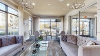 Located at 10051 Perimeter Pkwy in Charlotte, NC, this single-floor space functions as a sophisticated communal lounge designed to accommodate both relaxed gatherings and casual meetings. The interior is characterized by a warm, neutral color palette of taupe and greige tones, complemented by hardwood-style flooring and a patterned area rug that anchors the seating arrangements.

Large floor-to-ceiling windows and expansive glass panel walls line the perimeter, flooding the space with abundant natural light and providing open views of the surrounding landscaped exterior. Sheer white drapery frames the windows, softening the light and adding an elegant touch. Geometric chandelier pendants in a brushed metallic finish are suspended from the ceiling, enhancing the upscale ambiance.

The lounge area features tufted gray sofas with decorative accent pillows, glass-top coffee tables, and multiple round glass dining tables paired with upholstered medallion-back chairs, offering versatile seating options. Decorative wall art and sculptural wall-mounted orb clusters serve as distinctive design accents throughout.

A built-in beverage station, framed by textured mosaic columns, provides a functional amenity within the space. The overall design is polished and inviting, balancing contemporary aesthetics with comfortable functionality.