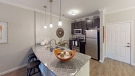 This single-floor residence offers a thoughtfully designed layout encompassing a living room, dining area, kitchen, two bedrooms, and two bathrooms. The living room is a welcoming space finished with crown molding, wood-style flooring, and natural light streaming through large windows with views of greenery. A glass door provides direct outdoor access, enhancing the open feel of the room.

The kitchen is well-equipped with dark espresso cabinetry, granite countertops, stainless steel appliances including a refrigerator and range, and a peninsula with pendant lighting overhead—ideal for both cooking and casual dining. Adjacent to the kitchen, the dedicated dining area features crown molding, wood-style flooring, and a chandelier overhead.

Both bedrooms offer plush carpeting, ceiling fans, and natural light from well-positioned windows. Each room provides a comfortable, restful atmosphere with ample space.

The two bathrooms are finished with neutral tile flooring. One features a tub-shower combination with subway tile surround and a curved curtain rod, while the second is a longer, corridor-style bathroom with a decorative shelf and tiled floors, adding a refined touch to both functional spaces.