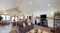 Located at 6614 North Cedar Road in Spokane, Washington, this single-story community clubhouse offers a welcoming and well-appointed gathering space designed for comfort and functionality. The centerpiece of the building is a generous great room characterized by soaring vaulted ceilings with recessed lighting, creating an open and airy atmosphere. Large wood-trimmed windows — including clerestory windows positioned high on the walls — flood the space with abundant natural light. The great room features plush carpeting and is arranged with comfortable seating areas anchored by a fireplace and flat-screen television, making it ideal for relaxed gatherings. A built-in display cabinet with glass-front doors serves as a stylish room divider between the lounge and entry areas, which are finished with neutral tile flooring. The kitchen is fully equipped with stainless steel appliances, including a refrigerator, electric range, microwave, and dishwasher, complemented by warm honey-toned cabinetry and tile countertops. French doors in the kitchen open directly to an outdoor patio, enhancing the connection to the exterior. A separate nook area provides a casual dining or activity space with its own window views, and an adjacent private office with carpeted flooring adds further versatility to the layout.