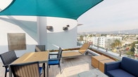 This rooftop terrace is a well-appointed outdoor amenity space positioned high above a bustling urban neighborhood, delivering panoramic views of the surrounding cityscape, palm tree-lined streets, and distant mountain ranges. The terrace is designed with distinct functional zones across its open-air layout, connected by light-toned porcelain tile flooring and dark wood-look tile walkways that lend a clean, contemporary aesthetic throughout.

A prominent lounge area features a deep-cushioned navy sectional sofa paired with a glass-top fire pit table, all sheltered beneath vibrant teal triangular shade sails that provide relief from the sun while adding a bold architectural accent. Floor-to-ceiling glass panel railings along the perimeter allow for unobstructed sightlines across the city skyline, maintaining a seamless connection to the expansive views.

An outdoor dining area includes a teak-style table with coordinating chairs, positioned alongside a reclining sun lounger — ideal for relaxing and enjoying the elevated vantage point. Raised planter beds integrated into the terrace walls and freestanding planters support greenery and low-growing vegetation, including a living green roof element that adds a sustainable, organic touch to the design. Wall-mounted speakers and security fixtures are discreetly placed throughout, completing this thoughtfully designed rooftop retreat.