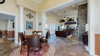 This single-floor community clubhouse or leasing center is designed with a classic, upscale aesthetic that creates a warm and inviting atmosphere. The space is anchored by soaring ceilings adorned with crown molding, and dramatic arched transom windows that allow generous natural light to pour in throughout. The main entry foyer greets visitors with large-format stone tile flooring in rich, multi-toned hues, white decorative columns, and two-tone wainscoting walls that lend a polished, traditional character.

The open lounge area serves as a central gathering space, furnished with rich leather sofas and chairs arranged around a glass-top coffee table. A floor-to-ceiling stacked stone fireplace with a white mantel and wall-mounted flat-screen television serves as a striking focal point. A ceiling fan overhead and arched windows add to the comfortable ambiance.

Adjacent to the lounge, a dedicated dining area features a round table with dark wood and leather chairs, positioned atop a decorative area rug. Framed artwork and ornamental ironwork wall décor add character throughout. The space also includes a fully equipped kitchenette with white cabinetry, dark countertops, and modern appliances. A private office area provides a functional workspace. French doors throughout connect the interior to the outdoors, further enhancing the bright and open feel of this well-appointed space.
