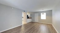 Located at 2415 West, this single-floor home offers a clean, functional layout with a welcoming open-concept design. The main living area is finished with durable hardwood-style laminate flooring in a warm, neutral tone that extends throughout the space, creating a cohesive and easy-to-maintain surface underfoot. Light gray walls complement the flooring and contribute to a bright, airy atmosphere.

A pendant light fixture hangs near the entry, adding a subtle decorative touch above what appears to serve as a dining area. Natural light enters through a window beside the front door, keeping the space feeling open and well-lit. The front entry door features gold-tone hardware, offering a small but deliberate design accent.

An open passthrough connects the living area to the kitchen, which is equipped with white upper cabinetry and rich, darker countertops that provide a pleasant contrast. A black appliance is visible in the kitchen, adding to the functional character of the cooking space. Adjacent to the kitchen, additional cabinetry and counter space are visible, suggesting a practical and organized layout.

Ceiling-mounted HVAC vents are present, ensuring climate control throughout. The overall design is clean and straightforward, making the space move-in ready and well-suited for everyday living.