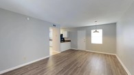 Located at 2415 West, this single-floor home offers a clean, functional layout with a welcoming open-concept design. The main living area is finished with durable hardwood-style laminate flooring in a warm, neutral tone that extends throughout the space, creating a cohesive and easy-to-maintain surface underfoot. Light gray walls complement the flooring and contribute to a bright, airy atmosphere.

A pendant light fixture hangs near the entry, adding a subtle decorative touch above what appears to serve as a dining area. Natural light enters through a window beside the front door, keeping the space feeling open and well-lit. The front entry door features gold-tone hardware, offering a small but deliberate design accent.

An open passthrough connects the living area to the kitchen, which is equipped with white upper cabinetry and rich, darker countertops that provide a pleasant contrast. A black appliance is visible in the kitchen, adding to the functional character of the cooking space. Adjacent to the kitchen, additional cabinetry and counter space are visible, suggesting a practical and organized layout.

Ceiling-mounted HVAC vents are present, ensuring climate control throughout. The overall design is clean and straightforward, making the space move-in ready and well-suited for everyday living.