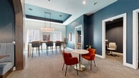 Located at 24720 Morton Ranch Rd in Katy, TX, this thoughtfully designed single-floor space offers a seamless blend of functionality and contemporary style. The interior is anchored by a bold teal and navy color palette, complemented by warm wood accents and plush carpeted flooring throughout.

The open living and dining areas form the heart of the space, where a tray ceiling with pendant lighting creates an elevated atmosphere. Vibrant burnt-orange velvet chairs surround a marble-topped bistro table, while a long bar-height communal dining table with upholstered seating sits beneath natural light from large windows.

A distinctive built-in entertainment wall features teal cabinetry with brass hardware, warm wood shelving, and a wall-mounted display — a standout architectural element that adds both character and utility. An elegantly arched alcove houses a cozy banquette dining nook with channel-tufted seating and a rich wood table, offering an intimate setting within the larger space.

Private rooms include a focused workspace with a built-in wood desk, task lighting, and textured dark wallcovering for a calm, distraction-free environment. A separate meeting room offers a round conference table, styled chairs, and a wall-mounted screen. The overall ambiance is modern yet inviting, balancing professional functionality with warm, design-forward aesthetics.