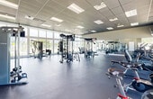 Located in Princeton, FL, this fitness center offers a generously sized, single-floor workout facility designed to support a full range of exercise needs. The space is defined by its striking floor-to-ceiling windows that line the walls, flooding the interior with abundant natural light and providing open views of the surrounding greenery and an adjacent outdoor pool area. The high drop-tile ceiling enhances the sense of vertical space, contributing to an airy and energizing atmosphere throughout.

The gym floor is finished with durable, cushioned flooring ideal for high-impact training and is organized to accommodate a wide variety of workout styles. Strength training equipment includes a multi-station cable machine, a Smith machine with an integrated squat rack, a dedicated free weight area with a dumbbell rack, and weight benches. Cardio offerings include treadmills, ellipticals, and stationary cycling bikes, all positioned to take advantage of the outdoor views. Wall-to-wall mirrors along one side of the facility visually expand the space and support proper form during workouts. Mounted flat-screen televisions provide entertainment throughout. The layout is open and well-organized, creating a clean, professional environment suitable for users of all fitness levels.