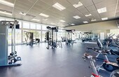 Located in Princeton, FL, this fitness center offers a generously sized, single-floor workout facility designed to support a full range of exercise needs. The space is defined by its striking floor-to-ceiling windows that line the walls, flooding the interior with abundant natural light and providing open views of the surrounding greenery and an adjacent outdoor pool area. The high drop-tile ceiling enhances the sense of vertical space, contributing to an airy and energizing atmosphere throughout.

The gym floor is finished with durable, cushioned flooring ideal for high-impact training and is organized to accommodate a wide variety of workout styles. Strength training equipment includes a multi-station cable machine, a Smith machine with an integrated squat rack, a dedicated free weight area with a dumbbell rack, and weight benches. Cardio offerings include treadmills, ellipticals, and stationary cycling bikes, all positioned to take advantage of the outdoor views. Wall-to-wall mirrors along one side of the facility visually expand the space and support proper form during workouts. Mounted flat-screen televisions provide entertainment throughout. The layout is open and well-organized, creating a clean, professional environment suitable for users of all fitness levels.