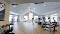 Located at 203 Magnolia Blvd in Port Wentworth, Georgia, this single-floor fitness center is thoughtfully designed to support a well-rounded workout experience. The space is defined by its striking vaulted ceiling, which rises to an impressive peak and is complemented by a combination of recessed lighting and a ceiling fan, creating a bright and comfortable environment. Durable hardwood-style flooring runs throughout, offering both visual warmth and a practical surface for exercise. Large windows line the walls, flooding the interior with natural light and contributing to the open, airy ambiance of the room. The fitness center is well-equipped with a diverse selection of cardio machines, including treadmills, ellipticals, stationary bikes, and a rowing machine, alongside strength and multi-use training equipment. The layout provides ample space between machines, allowing for easy movement and a comfortable workout experience. Motivational wall art adds a personal touch to the clean, neutral-toned décor. The combination of architectural character, generous natural light, and comprehensive equipment makes this fitness center a functional and inviting space for residents and users alike.