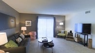 This single-floor space showcases a thoughtfully designed living room that balances contemporary style with comfortable functionality. The room features a bold charcoal accent wall that creates a striking contrast against the lighter surrounding walls and neutral-toned carpeted flooring, lending the space a modern, curated aesthetic.

A large sliding glass door or oversized window fitted with vertical blinds and complementary navy drape panels serves as the room's focal point, drawing in abundant natural light and offering potential access to an outdoor area. The bright influx of light enhances the open, airy feel of the space.

The living area is furnished with a dark upholstered sofa, a round tiered coffee table, a vibrant patterned armchair, and a green accent chair, together creating an eclectic yet cohesive seating arrangement. Floor and table lamps provide warm supplemental lighting. Wall art and decorative accessories add personality throughout. The carpeted flooring contributes to a soft, welcoming atmosphere, making this living space both visually appealing and practically comfortable for everyday living.