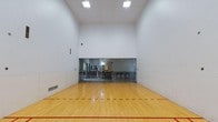 This dedicated racquetball court is a single-floor facility designed for serious play and recreation. The court is defined by its towering ceiling height, which provides ample vertical clearance essential for the fast-paced nature of the sport. The floor is finished in a high-gloss polished hardwood, with clearly marked red boundary and service lines that meet regulation standards. The expansive white walls on all sides offer clean, consistent playing surfaces, ensuring optimal ball rebound and visibility during play. A standout feature of the court is the full-width glass back wall, which allows spectators to observe matches from an adjacent viewing area without interrupting play. This viewing area is equipped with seating and provides a clear sightline across the entire court. Recessed floor vents along the base of the walls support climate control within the enclosed space, maintaining comfortable playing conditions. Overhead, flush-mounted rectangular lighting panels illuminate the court evenly, minimizing shadows and glare. The overall design is clean, functional, and purpose-built, making this a well-equipped space for both casual players and competitive athletes alike.