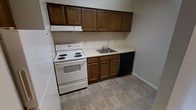 This single-floor space centers around a practical, efficiently arranged kitchen designed to maximize functionality within a compact footprint. The kitchen features a run of dark wood cabinetry that extends along one wall, with upper and lower cabinets providing ample storage for cookware and pantry essentials. The cabinet doors display a classic raised-panel style, lending a traditional character to the space.

Countertops in a light, neutral tone run the length of the lower cabinets, offering a clean work surface. A stainless steel single-basin sink is integrated into the countertop, accompanied by a standard faucet. Adjacent to the sink, a black built-in dishwasher provides added convenience.

The cooking area is equipped with a white electric coil range with a matching oven below, complemented by an overhead ventilation hood for managing cooking odors and steam. The flooring consists of tile in a muted, earthy tone with subtle variation, contributing to the space's low-maintenance, durable character.

The walls are finished in a light neutral color, and the overall layout follows a single-wall galley-style configuration, making efficient use of the available space. The kitchen presents a clean, move-in ready condition.