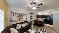 This single-story residence showcases an open-concept layout that connects the living area and kitchen, creating a spacious and cohesive environment well-suited for both relaxing and entertaining. The living area is anchored by a plush, dark sofa accented with decorative pillows, complemented by a bold patterned area rug and contemporary wall art that adds a pop of color and visual interest. A multi-light ceiling fan serves as a functional centerpiece overhead, providing both illumination and airflow throughout the space. A sleek floor lamp and a decorative corner shelving unit contribute additional style and storage. The open kitchen is visible from the living area and features dark cabinetry that contrasts warmly with lighter countertops. A breakfast bar with seating for multiple guests bridges the kitchen and living space, making it an ideal spot for casual dining or conversation. Bar-height chairs in deep red add a rich accent to the neutral palette. Black appliances and recessed lighting complete the kitchen's polished, contemporary look. The overall ambiance is warm and modern, with a cohesive dark-toned color scheme balanced by lighter walls and flooring throughout.