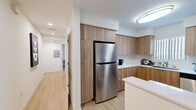 This single-floor residence offers a well-planned layout that maximizes every square foot across six distinct spaces: a bedroom, bathroom, kitchen, hallway, closet, and laundry area. The kitchen is a standout feature, showcasing warm wood-tone cabinetry that extends floor to ceiling, white quartz-style countertops, and a full suite of stainless steel appliances including a gas range, dishwasher, over-the-range microwave, and refrigerator. A built-in peninsula provides additional counter space and a natural division between the kitchen and the rest of the unit. Light wood-look flooring runs consistently throughout, lending warmth and visual continuity to the home. The bedroom is a comfortable, private retreat with a built-in wall unit that integrates cabinetry, a media center, and a dedicated refrigerator, reflecting clever, purpose-driven design. The bathroom is clean and contemporary, featuring a wall-mounted sink, a backlit vanity mirror, and a full bathtub with a sliding glass shower enclosure. Recessed lighting and flush-mount ceiling fixtures keep the space bright and functional. The hallway connects all areas efficiently, with an electrical panel neatly tucked along the wall. Overall, the residence presents a cohesive, modern aesthetic with practical amenities suited for comfortable everyday living.