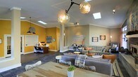 This single-floor space showcases a thoughtfully designed open-plan interior that seamlessly blends functional and social zones. The layout incorporates a dedicated workspace area, distinguished by a bold mustard-yellow accent wall that adds a vibrant, energetic contrast to the otherwise neutral taupe and white color palette throughout the rest of the space.

Natural light is a standout feature, provided by multiple skylights embedded in the vaulted ceiling, which floods the interior with brightness and enhances the sense of openness. A striking sculptural chandelier, featuring clustered globe pendants on an industrial-style branching frame, serves as a dramatic focal point overhead.

The lounge area is furnished with generous grey sofas accented with colorful throw pillows and plaid blankets, arranged around a wood-topped coffee table. A natural wood communal table with contemporary sculpted chairs occupies the foreground, suggesting a collaborative or dining function.

Artwork and decorative pieces are displayed throughout, adding character and personality to the walls. Dark hardwood flooring grounds the space and ties the different zones together, contributing to the warm, eclectic, and welcoming ambiance overall.