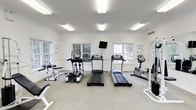 This single-floor fitness room offers a clean, functional workout environment designed to accommodate a variety of exercise routines. The space is finished with neutral white walls accented by crown molding, lending a polished appearance to the room. Carpeted flooring runs throughout, providing a comfortable surface underfoot.

Natural light flows in through multiple double-pane windows along the walls, offering views of outdoor greenery and keeping the space bright and inviting. Overhead, rectangular fluorescent light fixtures provide consistent, even illumination throughout the room.

The cardio equipment lineup includes two treadmills and an elliptical trainer positioned near the windows, alongside an upright stationary bike. A wall-mounted flat-screen television is centrally positioned above the windows, allowing users to watch while exercising. Additional strength and flexibility equipment, including a weight bench and a rack, are arranged along the sides of the room.

Informational fitness posters are displayed on the walls, adding an educational element to the space. The overall layout is open and uncluttered, ensuring ease of movement between equipment and creating a comfortable, welcoming atmosphere for users of all fitness levels.
