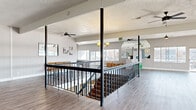 This expansive single-floor commercial space offers a versatile and open layout designed to accommodate a variety of uses. Light wood laminate flooring runs throughout, lending a clean and modern aesthetic to each area. The central feature of the space is a wide staircase framed by elegant black iron railings with turned balusters and structural steel columns, creating a striking visual anchor point.

The main open area is generously proportioned, bathed in natural light from large windows along multiple walls that offer views of the surrounding neighborhood. Multiple ceiling fans are distributed throughout, ensuring comfortable airflow across the broad floor plan. A recreational area includes a pool table positioned beneath a ceiling fan and alongside floor-to-ceiling sliding glass doors and windows.

A secondary seating area features classic wooden bench pews, evoking a gathering or community atmosphere. One section of the space includes a white brick fireplace surround, adding a warm architectural detail, along with a glass-paneled interior door. Large mirrors are integrated into certain walls, visually expanding the space further. A service counter or reception area is visible near the windows at one end. The overall ambiance is bright, open, and adaptable, well-suited for community programming, events, or organizational use.