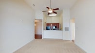This single-floor residence is defined by its impressive vaulted ceilings that lend a sense of openness and grandeur to the living area. The main living space features soft, neutral-toned carpet underfoot and crisp white walls that reflect light throughout, creating a bright and welcoming atmosphere. A ceiling fan with an integrated light fixture is centrally mounted, providing both comfort and functional illumination.

The open-concept design connects the living area directly to the kitchen via a raised pass-through counter, enhancing the sense of flow and connectivity throughout the space. The kitchen is equipped with rich, dark wood cabinetry that provides a warm contrast to the light surroundings, complemented by dark countertops, a black refrigerator, and a pendant light fixture that adds a decorative touch over the counter.

An adjacent area, visible through an open doorway, features hardwood-style flooring and an overhead light fixture, suggesting a dining area or secondary room that extends the functional living space. Additional architectural details include a built-in wall unit and clear sightlines across multiple zones, making the layout feel cohesive and well-organized. Overall, the space offers a clean, comfortable, and efficiently designed interior with a warm, understated aesthetic.